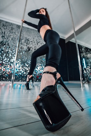 Sexy woman in high heels dancing with pylon in art-studio. Poledance on shining wall background.