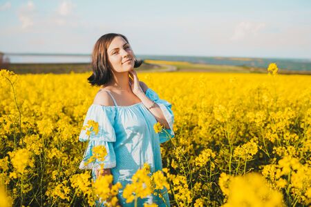 Portrait of young pretty woman in blue dress posing in rapeseed yellow flowers field. Spring time,...