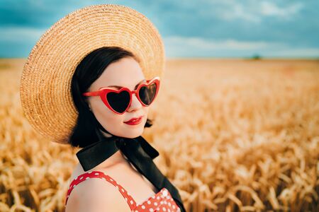 Portrait of stylish woman in wheat field. Old fashioned girl in straw hat, heart-shaped eyeglasses,...