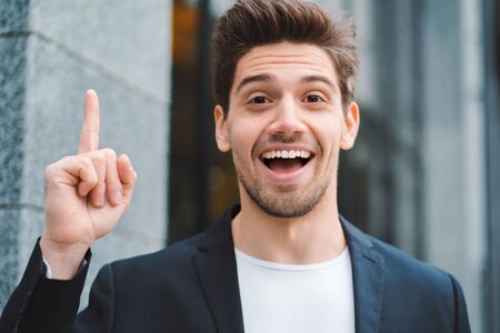 Portrait of young thinking pondering businessman having idea moment pointing finger up on office building background. Smiling happy student man showing eureka gesture.の写真素材