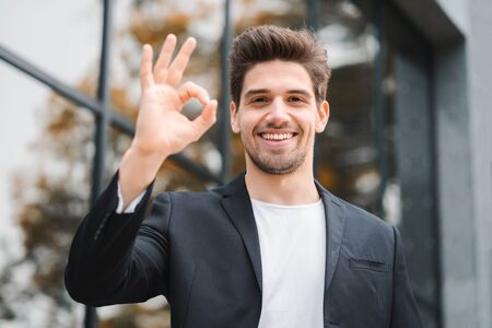 Successful happy man looking to camera and showing OK sign on office building background....