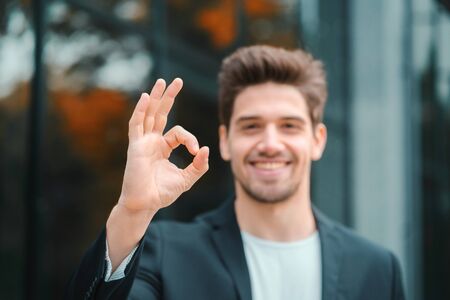 Successful happy man looking to camera and showing OK sign on office building background. Professional male manager wearing corporate suit jacket. Student guy outdoorsの写真素材