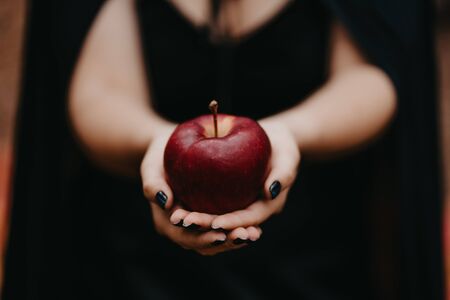 Black witch holding red apple in hands on autumn forest background. Horror, halloween, cosplay holiday, magic conceptの写真素材