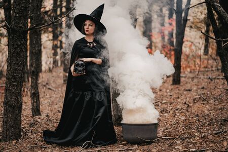 Black witch holding skull in hands, stroking it on autumn smoke forest background. Horror, halloween, cosplay holiday, magic conceptの写真素材