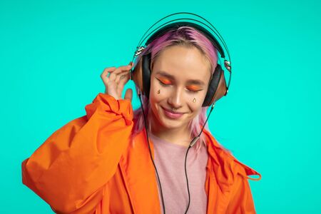 Pretty girl with pink hair enjoys listening to music. Hipster woman with extraordinary appearance in orange raincoat on blue background.の写真素材