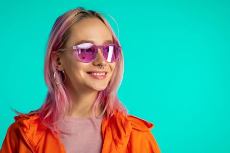 Cheerful odd girl in blue studio. Smiling woman with nose piercing, sunglasses. Vibrant colorful...