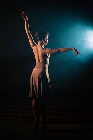Ballerina is practicing her moves on dark stage. Young girl dancing in beige dress, spinning around...