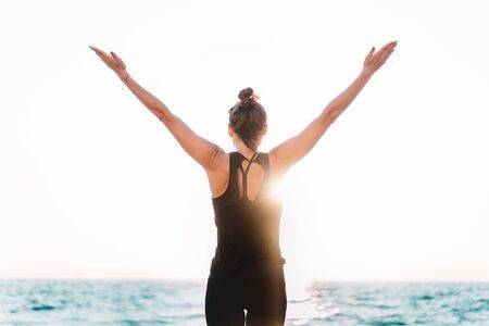 Young woman with open up hands to the sun, she practicing yoga exercises at sunrise light, minimalist scene. Doing asana. Healthy lifestyle concept.の写真素材