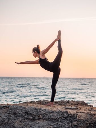 Young woman with curly hair in black costume practicing yoga at sunrise light, minimalist scene. Doing asana. Healthy lifestyle concept.の写真素材