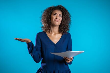 Serious african woman is unsatisfied with papers, bills, documents or reports on blue background. Young female office employee isolated in studio.の写真素材