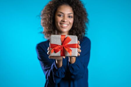 Mixed race woman gives gift and hands it to the camera. She is happy, smiling. Girl on blue...