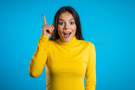 Smiling happy student girl showing eureka gesture. Portrait of young thinking pondering business woman having idea moment pointing finger up on blue backgroundの写真素材