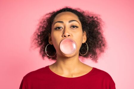 Playful girl in red wear blows bubblegum, chewing gum in slow motion. Pretty african american woman stands on pink backgroundの写真素材