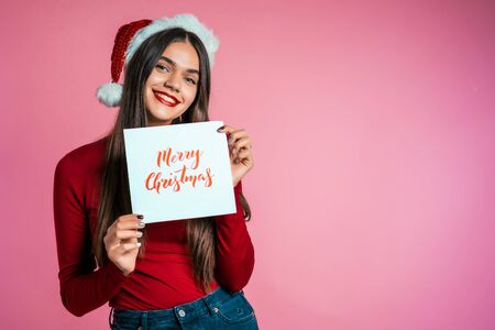Attractive girl in Santa hat with nameplate Merry Christmas. Portrait of lady on pink background. New year concept. Happy pretty woman smiling to camera.の写真素材