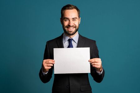 Businessman holding white horizontal a4 paper poster. Copy space. Smiling handsome man in corporate suit with tie on blue background.の写真素材