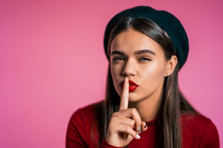 Beautiful woman with red lips holding finger on her lips over pink background. Gesture of shhh, secret, silence. Close up.の写真素材