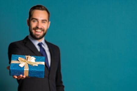 Handsome businessman in suit gives gift box and hands it to camera. He is happy, smiling. Man with beard on blue background. Positive holiday shot.の写真素材