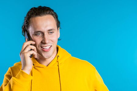 Young man in bright colorful yellow wear talking on mobile cell phone on blue background. Trendy guy have conversation. Smartphone, technology concept.の写真素材