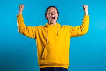 Yes winner gesture. Happy european man. Handsome guy with stylish hairdo surprised to camera over blue backgroundの写真素材