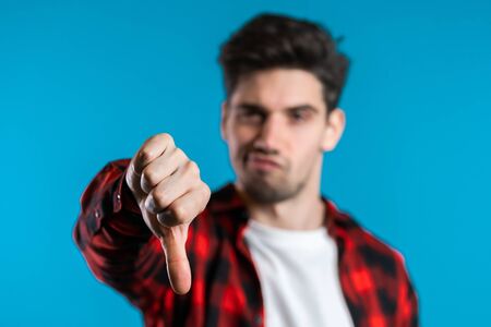 Young handsome man standing on blue studio background expressing discontent and showing thumb down...