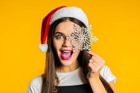 Beautiful woman in Santa hat holding big snowflake - symbol of winter. Studio yellow background.の写真素材