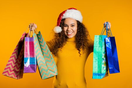 Happy young woman in Santa hat with colorful paper bags after shopping isolated on yellow studio background. Winter sale, purchases, spending money on gifts concept.の写真素材