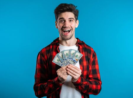 Man in red plaid shirt with surprised happy face holding US currency. Person with money. New dollars in hands on blue studio background .の写真素材