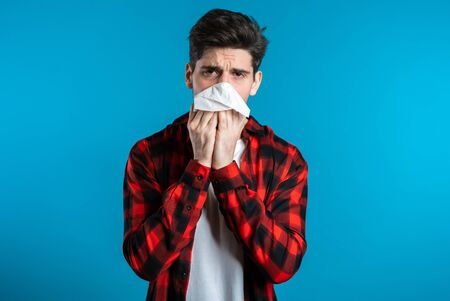 Young man sneezes into tissue. Isolated guy is sick, has a cold or allergic reaction. Health, medicine, illness, treatment conceptの写真素材