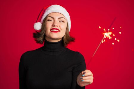 Trendy hipster girl in Santa hat with bengal sparkler fire on red background. Smiling, happy lady. Portrait of young pretty woman celebrating Christmas or New year party.の写真素材