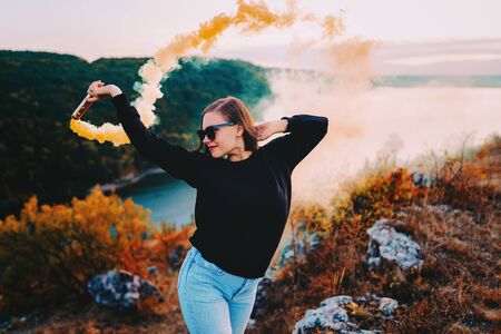 Young pretty woman with orange smoke bomb or grenade in dramatic moody lighting. Girl in black sweatshirt on a cliff above the riverの写真素材