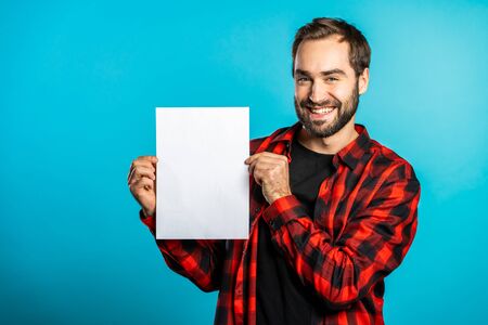 Handsome man holding vertical white a4 paper poster. Copy space. Smiling hipster guy in red plaid shirt on blue background.の写真素材