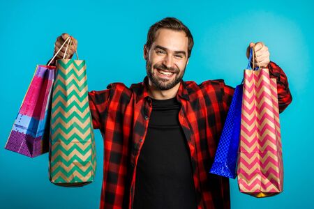 Handsome young man holds shopping paper bags on blue studio background. Guy bought presents on sales with discounts.の写真素材