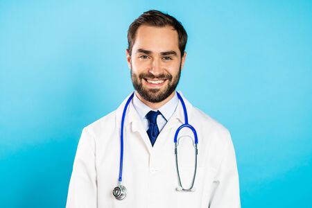 Portrait of young smiling man in professional medical white coat is isolated on blue studio background. Doctor with beard and stethoscopeの写真素材