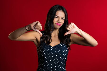 Young pretty woman standing on red studio background expressing discontent and showing thumb down gesture at camera. Portrait of girl with sign of dislikeの写真素材
