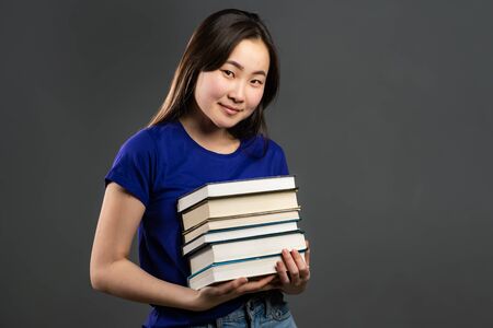 Asian student on grey background in studio holds stack of university books from library. Woman...