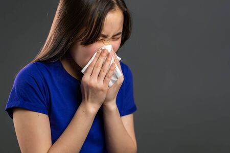 Young asian girl sneezes into tissue. Isolated woman on grey studio background is sick, has a cold or allergic reaction. Coronavirus, epidemic 2020, illness conceptの写真素材