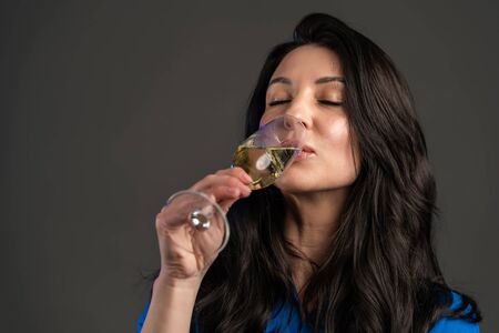 Attractive latin mature woman smiling and holding glass of champagne or wine on grey studio background. Drinking alcohol conceptの写真素材