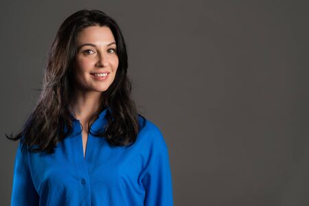 Charming mature woman isolated on grey studio background. Portrait of adult lady with long hair in blue shirt with smile looking to camera. Copy spaceの写真素材