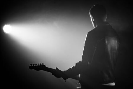 Rock guitarist man in leather jacket standing his back in smoky studio or stage masterfully playing electric guitar. View of unrecognizable musician in the spotlightの写真素材