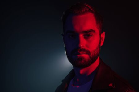 Portrait of young caucasian man with beard in leather jacket illuminated neon light looking at camera. Dark smoke studio background.の写真素材