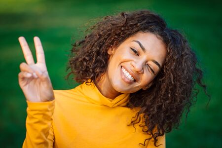 Portrait of african american girl showing peace, ok gesture in green park. Young woman with curly hairstyle in yellow wear enjoys nature.の写真素材