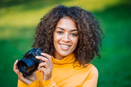 Young pretty african woman in yellow takes pictures with DSLR camera over green background. Girl smiling having fun as photographer.の写真素材