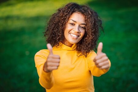Portrait of african american girl showing thumbs up like gesture in green park. Young woman with curly hairstyle in yellow wear enjoys nature.の写真素材