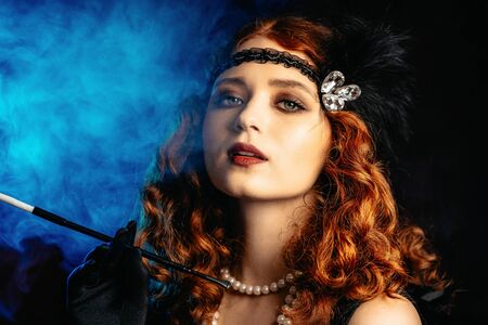 Old-fashioned woman dressed in style of Flappers posing with cigarette in mouthpiece on dark...