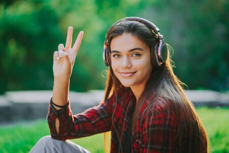 Young teenager listens to music through headphones in park.Girl in red plaid shirt smiles, dancing to rhythm.Concept of student life, freedom, modern youthの写真素材