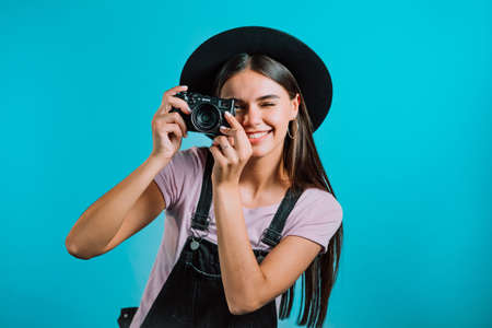Young pretty woman in overall takes pictures with DSLR camera over blue background in studio. Girl smiling and having fun as photographer.の写真素材