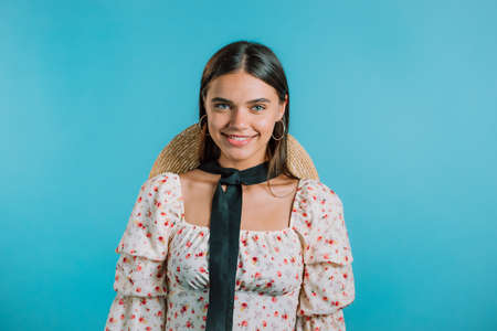 Young smiling woman with hat looking to camera. Portrait of cute beautiful girl in floral dress on blue studio wall background.の写真素材