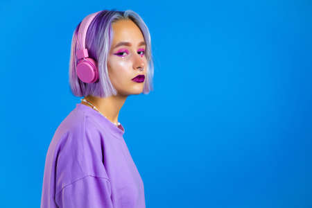 Pretty girl with dyed violet hair listening to music, smiling, dancing in headphones in studio against blue background. Music, dance, radio conceptの写真素材