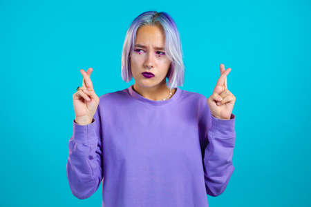 Pretty unusual teenager girl with dyed purple hair praying over blue background. Woman begging...