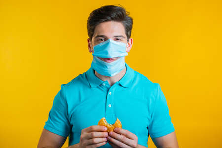 Young man in protective medical mask eating burger on yellow studio background. Guy makes fun, shows how to live during an epidemic.の写真素材